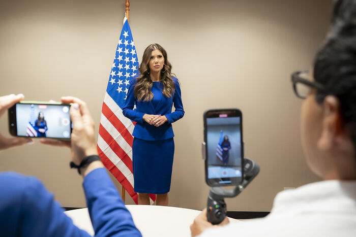 Woman standing by American flag being recorded on smartphones, related to Minneapolis immigration agents and body cameras.