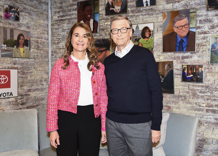 Bill Gates and Melinda French Gates posing together indoors with framed photos on brick wall behind them.