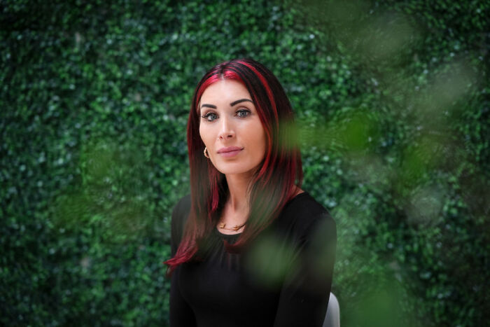 Laura Loomer posing with red highlights in hair, wearing black top against a green leafy background in a portrait photo.