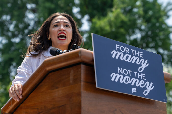AOC speaking passionately at a podium outdoors with a sign reading for the many not the money. AOC speaking passionately at a podium outdoors with a sign reading for the many not the money.