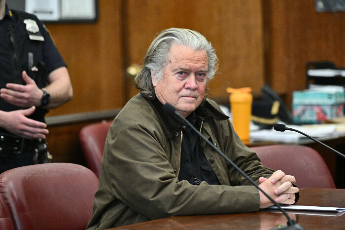 Steve Bannon sitting in a courtroom, wearing a green jacket, with microphones in front amid Epstein ties controversy.
