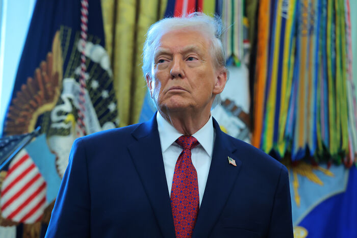 Donald Trump in a blue suit and red tie standing in the Oval Office with presidential flags and decor in the background