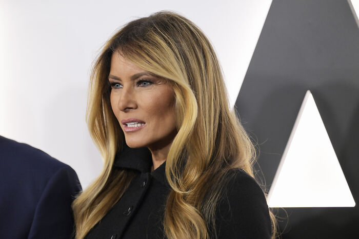 Melania Trump with serious expression, wearing black outfit, reacting during a public event with a geometric background.