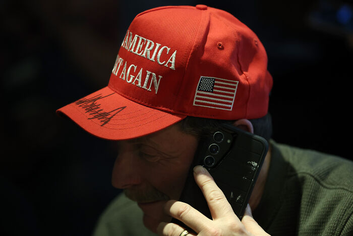 Man wearing red MAGA hat with American flag patch speaking on phone in low light, illustrating MAGA ideologies on dates. Man wearing red MAGA hat with American flag patch speaking on phone in low light, illustrating MAGA ideologies on dates.