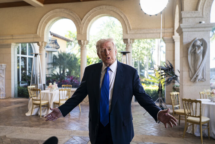 Donald Trump at an event, wearing a suit and blue tie, with an unusual appearance suggesting skipped makeup.