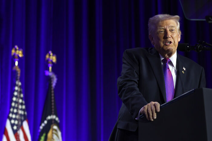 Donald Trump speaking at National Prayer Breakfast, delivering a lengthy speech with American and presidential flags in the background.
