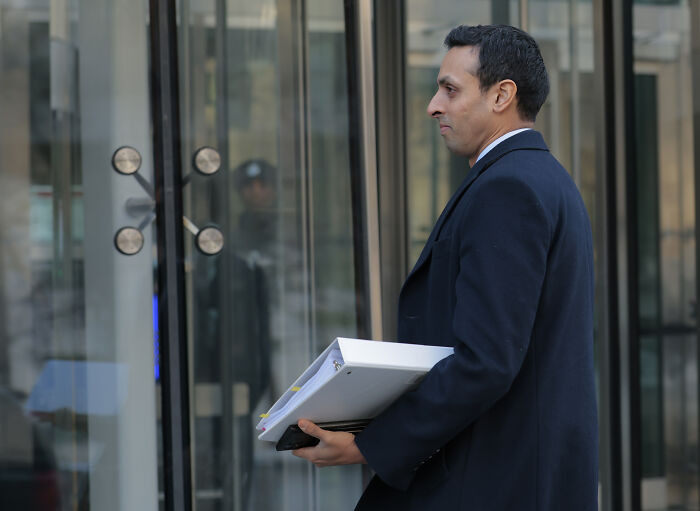 Man in dark coat holding documents, entering glass building, symbolizing reps turning up heat on King Charles scandal.