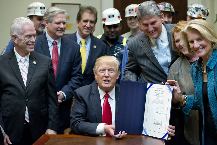 Donald Trump holding coal industry trophy surrounded by officials and workers wearing hard hats during coal event.