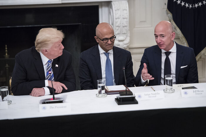 Jeff Bezos speaking at a business meeting with Donald Trump and Satya Nadella, highlighting Trump ties controversy.