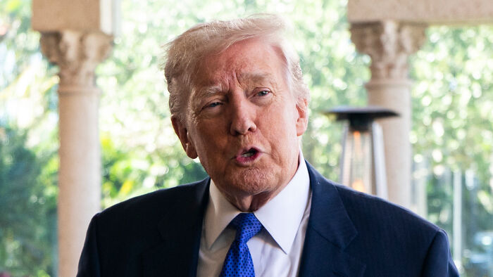 Donald Trump speaking at an outdoor event, wearing a dark suit and blue tie, without obvious makeup application visible.