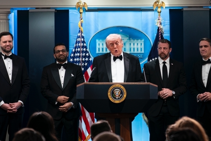 Trump officials in tuxedos, including Trump at a podium, speaking at a formal event, with the US flag in the background.
