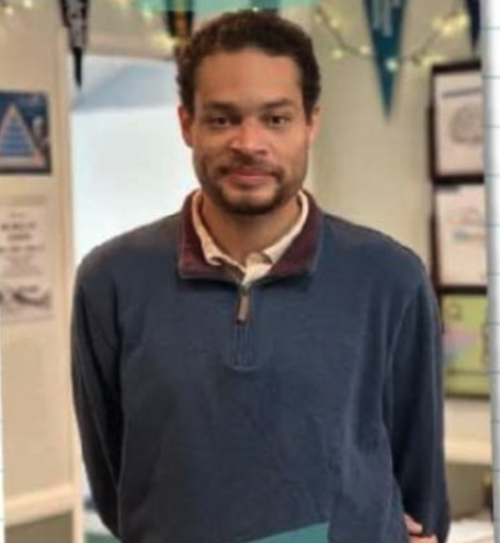 A medium close-up of the alleged WHCD shooter, a man with short curly hair and a beard, wearing a blue zip-up sweater.